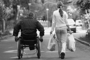 A black and white view from behind of a man in a black jacket pushing a wheelchair next to a woman in white who is walking and carrying grocery bags.
